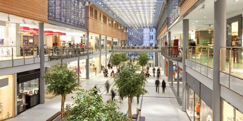 Image taken inside a shopping mall. There are lots of people walking around going to stores, buying things and consuming food and beverages. There are two floors with shops and boutiques. The ceiling is glass. The architecture is very modern and new and the general feel of the images is bright, clean, safe and positive.